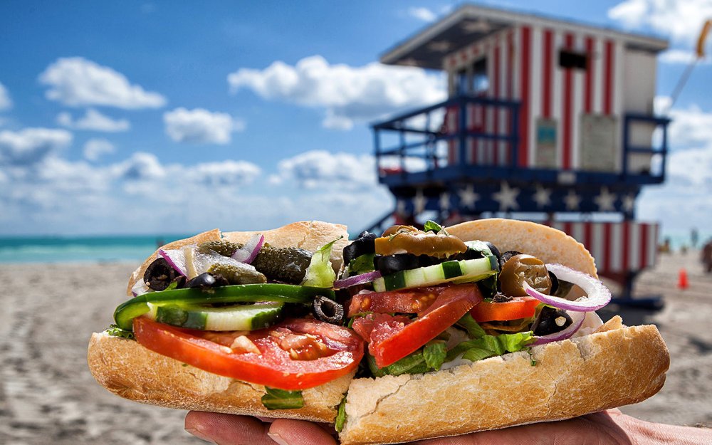 Mouthwatering sandwich overflowing with fresh ingredients and vibrant colors, served on a crispy baguette, beach in the background with lifeguard stand